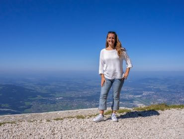 Silvia Spinnato al monte Untersberg_SchmitzbergerSabrina-2 Italiana nel Salisburghese sorride in cima a una montagna con alle spalle una valle panoramica e un cielo azzurro.