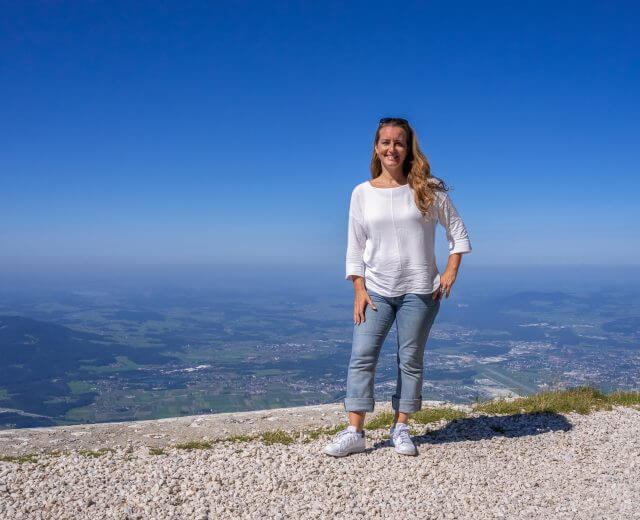 Italiana nel Salisburghese sorride in cima a una montagna con alle spalle una valle panoramica e un cielo azzurro.