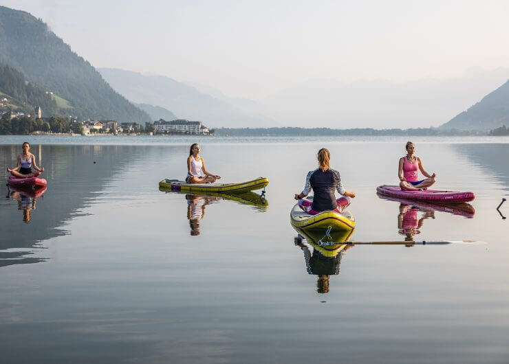 Quattro persone sedute su tavole da paddle fanno yoga su un lago calmo, circondato da montagne lontane.