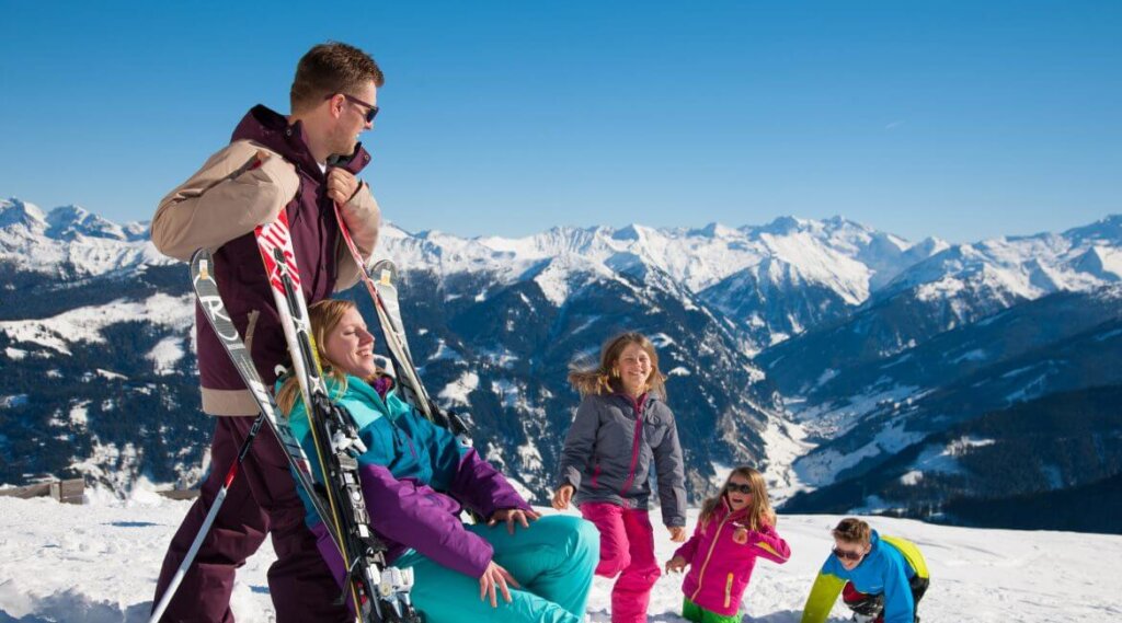 Famiglia sulla neve sotto il sole primaverile: genitori con sci, bambini che giocano, sullo sfondo un panorama alpino aperto.