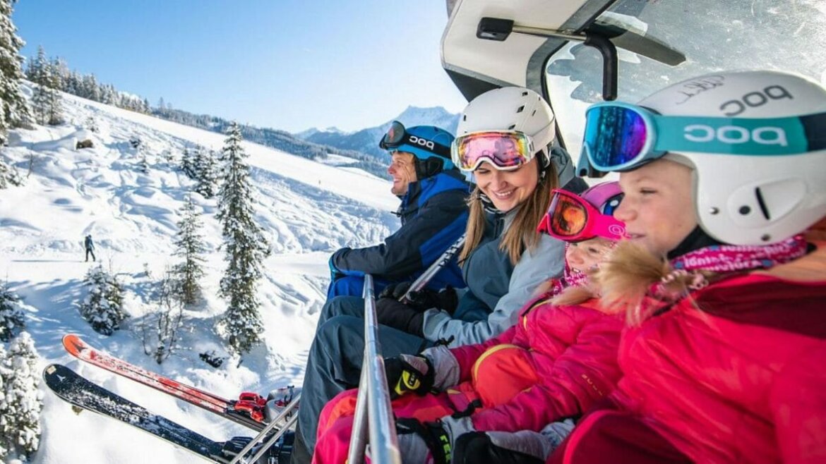 Famiglia su una seggiovia con cupola protettiva: bambini davanti, genitori dietro e panorama alpino sullo sfondo.