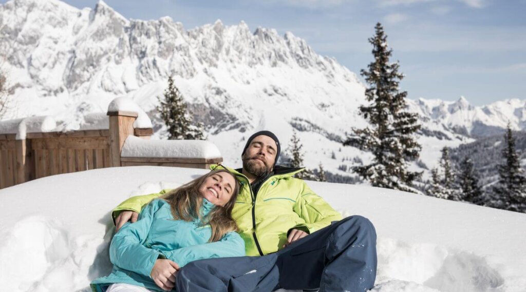 Coppia sdraiata sulla neve in abiti da sci, abbracciata e sorridente sotto il sole primaverile in montagna.