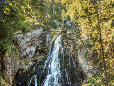 Una cascata scende da una parete rocciosa circondata da una fitta foresta verde e da colline lontane.