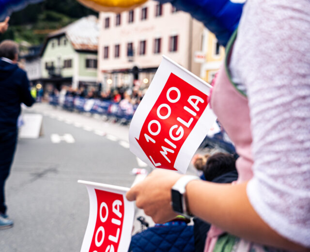 Persona che tiene in mano la bandiera della 1000 Miglia durante un evento di strada, con folla ed edifici sfocati sullo sfondo.