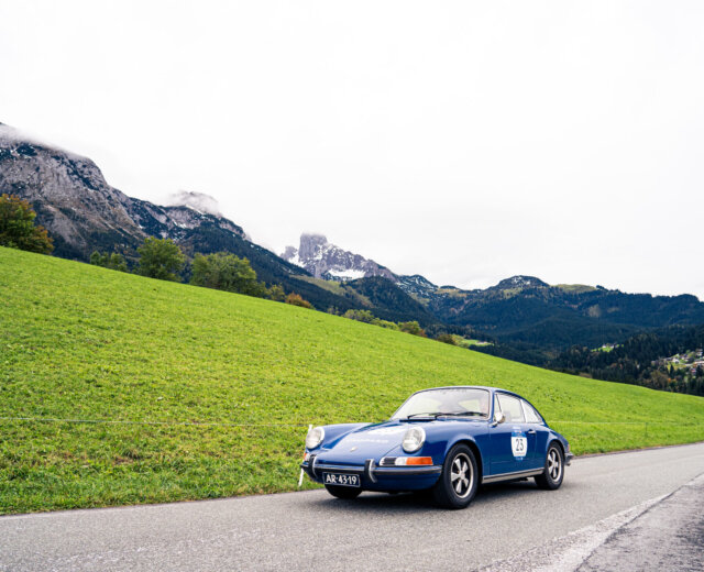 Un'auto d'epoca blu percorre una strada di montagna con colline verdi e cime rocciose sullo sfondo.