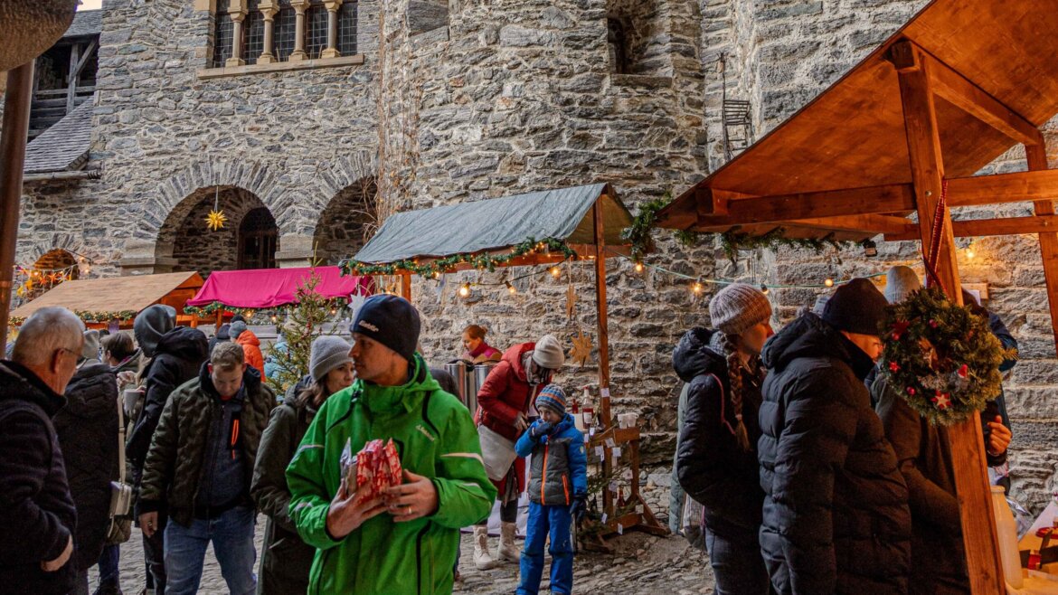 Persone si radunano attorno al mercatino di Natale davanti al castello di Finstergrün, sorseggiando vin brulé in un’accogliente atmosfera serale natalizia. (Vista ingrandita)