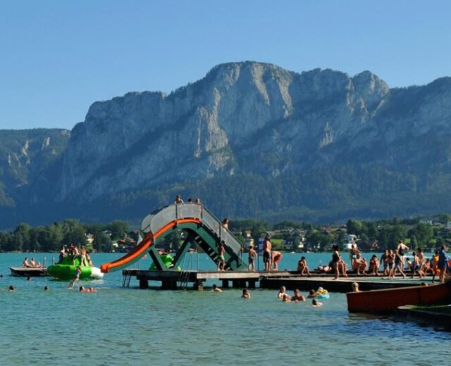 Persone sul pontile con scivolo dell’Aquapark dell’Alpenseebad di Mondsee con montagne sullo sfondo