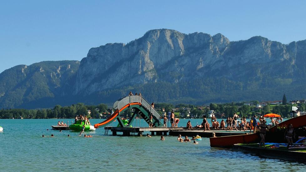 Persone sul pontile con scivolo dell’Aquapark dell’Alpenseebad di Mondsee con montagne sullo sfondo