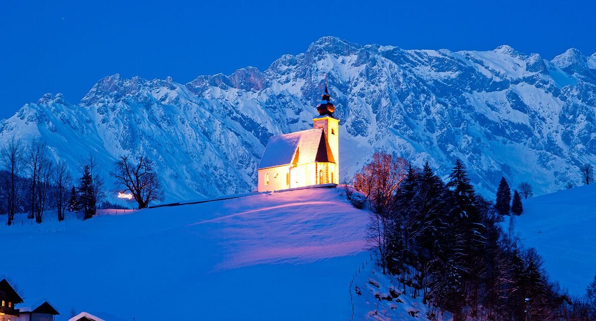 Chiesa di San Nicola illuminata sulla collina innevata con il massiccio dell’Hochkönig sullo sfondo (Vista ingrandita)