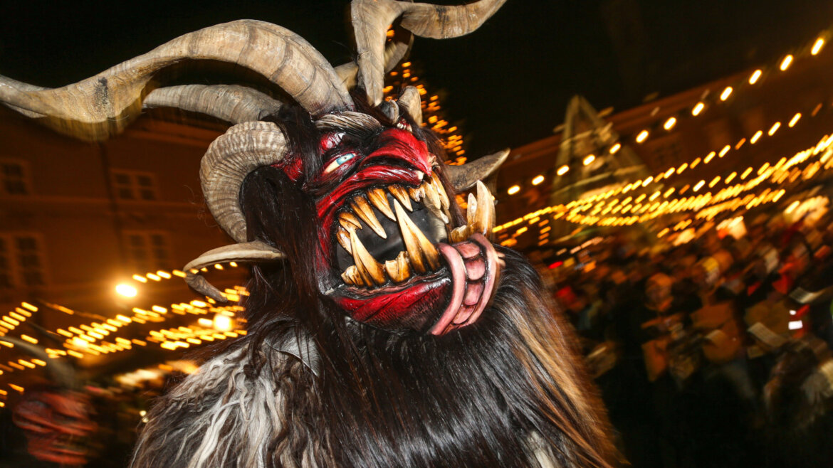 Diavoli alpini che accompagnano tradizionalmente San Nicola al Mercatino di Gesù Bambino di Salisburgo con la piramide di vetro che protegge la Colonna di Maria sullo sfondo (Vista ingrandita)