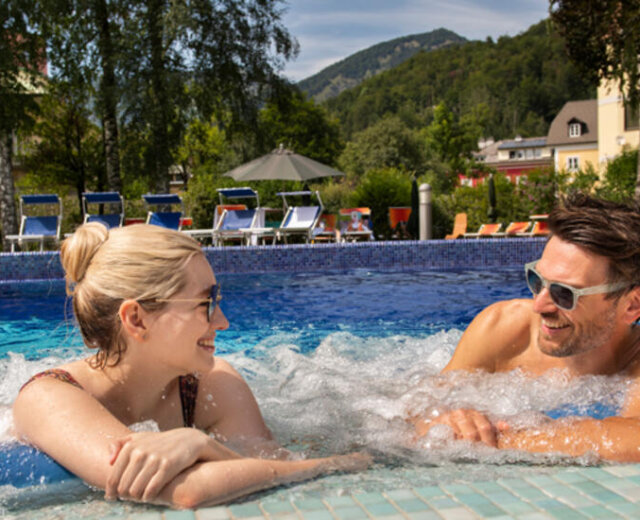 Due persone sorridono e si rilassano in una piscina con montagne e sedie a sdraio sullo sfondo.