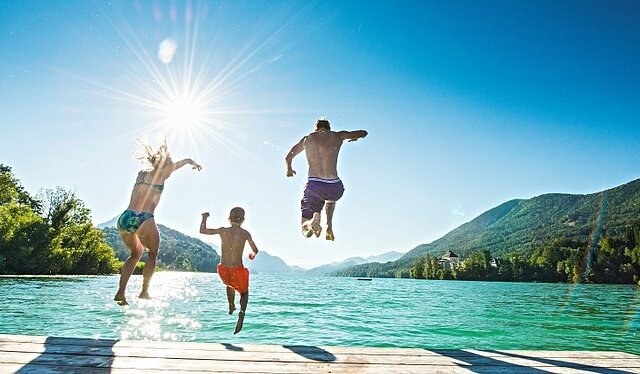 Tre persone saltano da un molo in un lago, con un cielo azzurro e montagne verdi sullo sfondo.