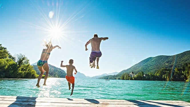 Tre persone saltano da un molo in un lago, con un cielo azzurro e montagne verdi sullo sfondo.