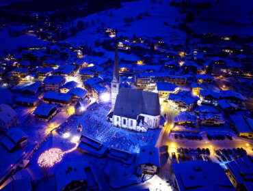 Veduta aerea di un villaggio innevato di notte, con le luci che illuminano la chiesa centrale e gli edifici circostanti.