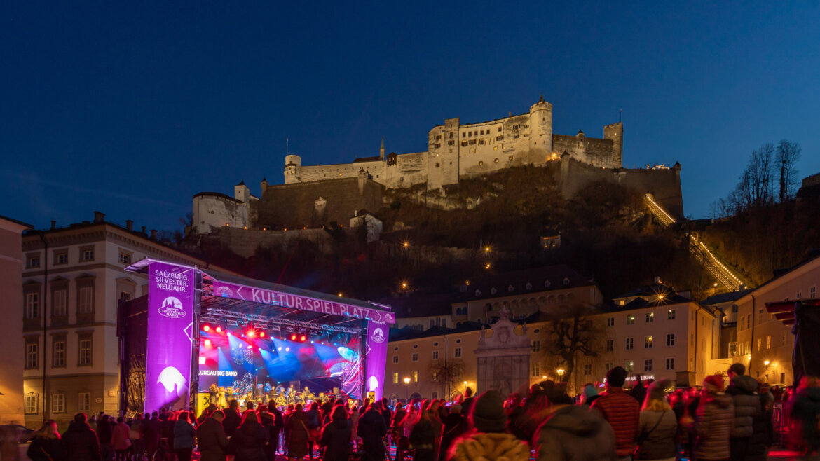 Folla davanti al palco illuminato in Piazza del Capitolo a Salisburgo con la fortezza di Hohensalzburg che domina il centro storico (Vista ingrandita)