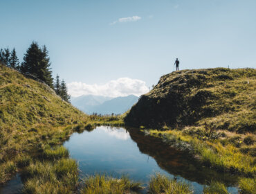Persona in piedi su una collina erbosa che si affaccia su un piccolo stagno con montagne e alberi sullo sfondo.