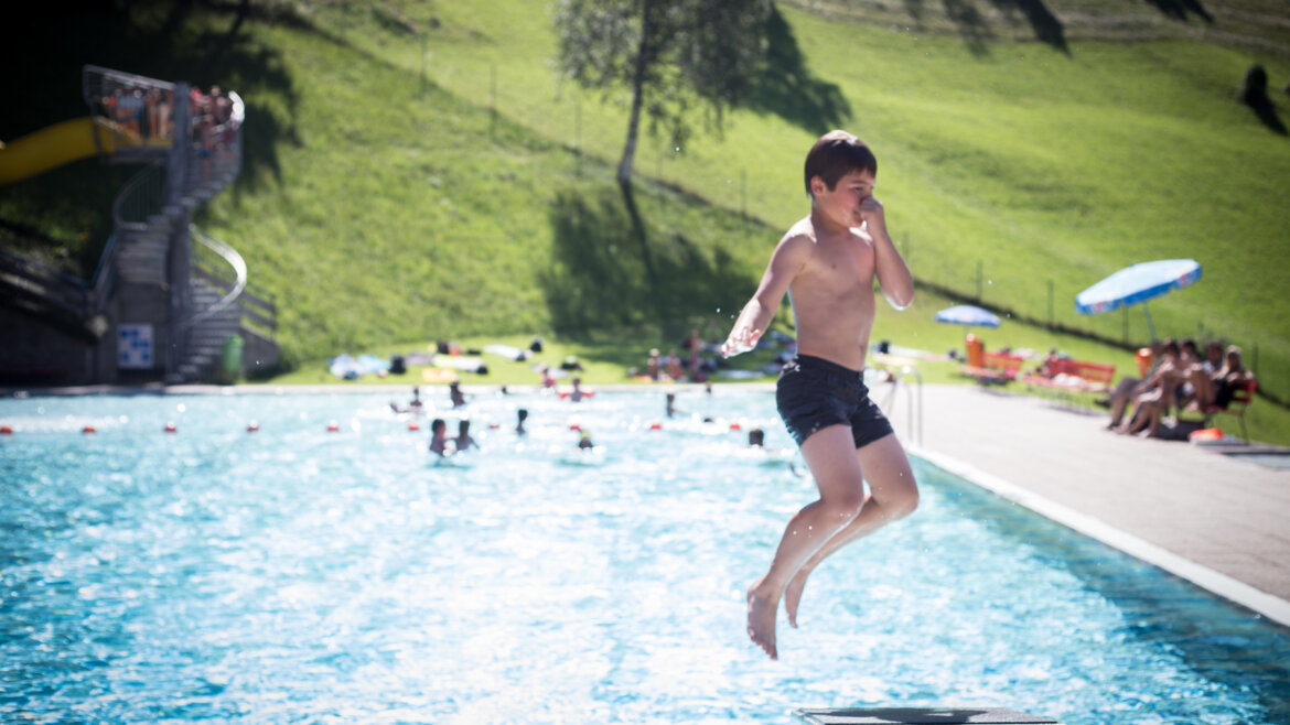 Un ragazzo in costume da bagno salta in una piscina all'aperto, tenendosi il naso, con persone e alberi sullo sfondo. (Vista ingrandita)