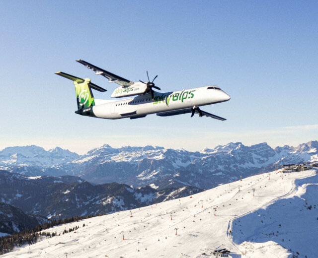 SkyAlps sopra un comprensorio sciistico alpino Un aereo SkyAlps sorvola un comprensorio sciistico alpino con piste innevate e impianti di risalita sotto un cielo limpido.