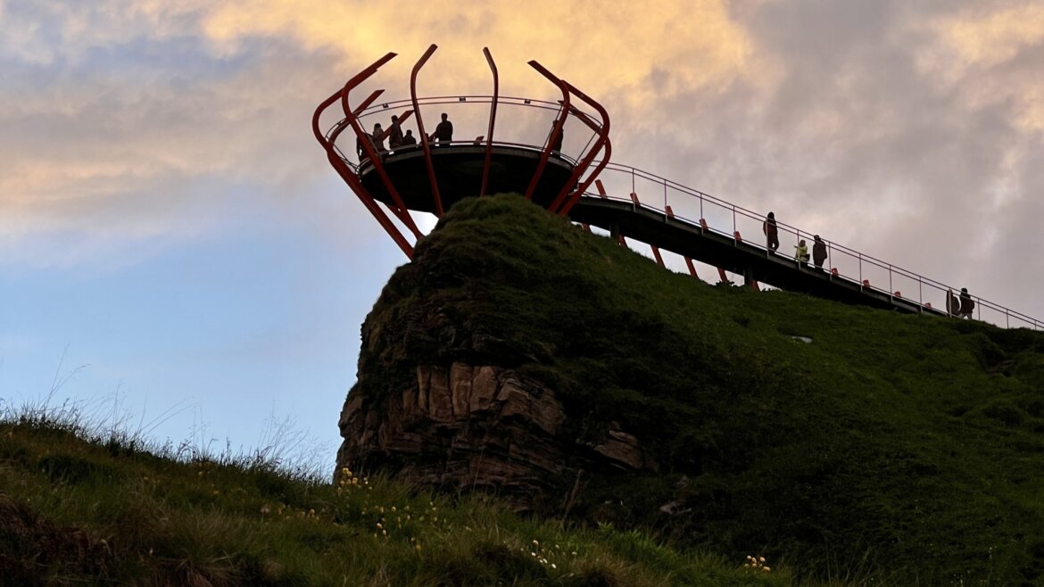 Proteso sul paesaggio alpino, il Glocknerblick sullo Stubnerkogel offre una vista privilegiata sulle montagne di Bad Gastein. © Azzurra Forti – BabyTrekking Al tramonto, alcune persone sulla piattaforma panoramica Glocknerblick sullo Stubnerkogel, costruita su uno sperone roccioso e collegata da una passerella, sotto un cielo nuvoloso. (Vista ingrandita)