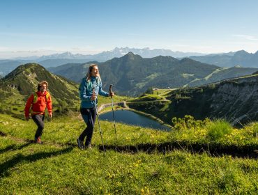 Twee mensen wandelen op het met gras begroeide bergpad van Sportwelt, met schilderachtige heuvels, een meer en verre bergtoppen onder een heldere hemel.