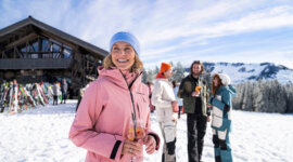 tussenstop in de hut ©SalzburgerLand Vrouw in roze jas geniet van après-ski drankje, glimlachend met vrienden bij een skihut in besneeuwde bergen.