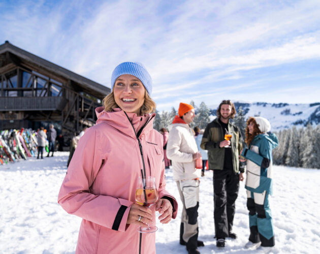 tussenstop in de hut ©SalzburgerLand Vrouw in roze jas geniet van après-ski drankje, glimlachend met vrienden bij een skihut in besneeuwde bergen.