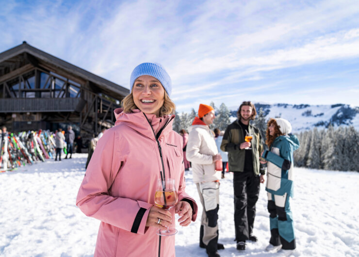 tussenstop in de hut ©SalzburgerLand Vrouw in roze jas geniet van après-ski drankje, glimlachend met vrienden bij een skihut in besneeuwde bergen.