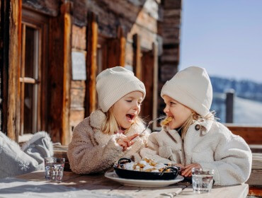 Kinder am Hochkönig Twee jonge kinderen in winterkleren delen een toetje buiten een rustieke houten hut - perfect voor Familietips.