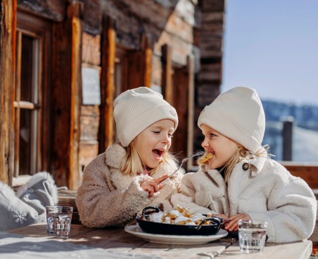 Twee jonge kinderen in winterkleren delen een toetje buiten een rustieke houten hut - perfect voor Familietips.
