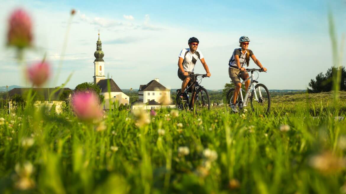 Pärchen beim Radfahren – Tauernradweg Twee mensen rijden over een van de mooiste fietsroutes door een grasveld, een dorp en een kerk op de achtergrond.