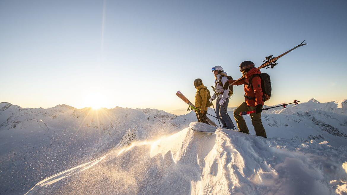 Drie skiërs staan bij zonsopgang op een besneeuwde bergtop, klaar voor een dag vol spannende wintersportavonturen.