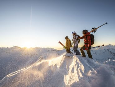 Drie skiërs staan bij zonsopgang op een besneeuwde bergtop, klaar voor een dag vol spannende wintersportavonturen.