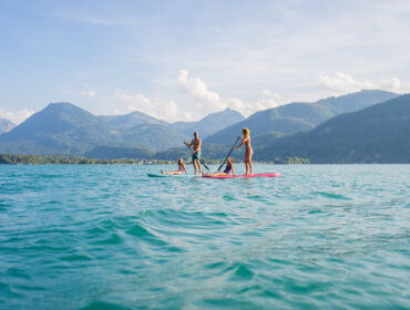 Zwemplezier aan de Wolfgangsee Vier mensen paddleboarden op een meer, glijden over het water met bergen en blauwe lucht op de achtergrond.