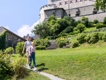 Een stel wandelt over een tuinpad onder een stenen kasteel, genietend van een zonnige dag met hun SalzburgerLand Card.