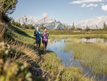 salzburger-gipfelspiel-der-gerzkopf-ein-fazinierender-berg-mit-sagenumwobenen-see-ein-mystisches-gipfelmoor-und-einer-wunschglocke-salzburger-sportwelt_michael-groessinger-2 Twee mensen trailrunning naast een bergmeer in de Sportwelt, met schilderachtige bergtoppen en blauwe lucht achter hen.