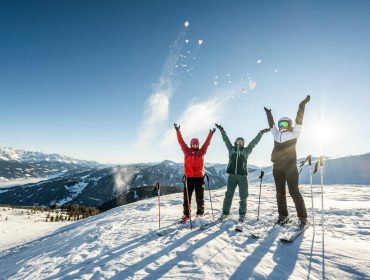 salzburger-sportwelt-c-lorenz-masser-153 Drie skiërs vieren feest op een besneeuwde Sportwelt-berg, de armen geheven onder de blauwe hemel en het zonlicht.