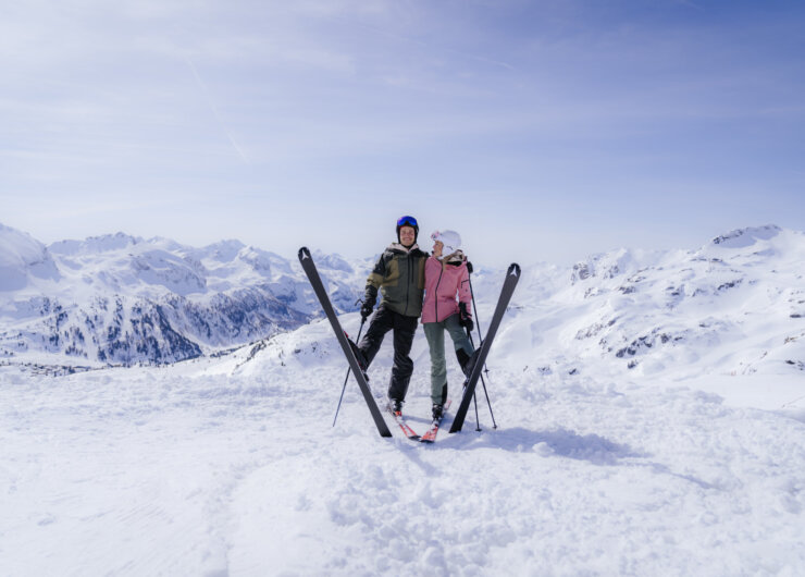 ski_alpin_SalzburgerLand_Wintermood_Pärchen-3 Twee skiërs die op een besneeuwde bergtop staan met hun ski’s gekruist.
