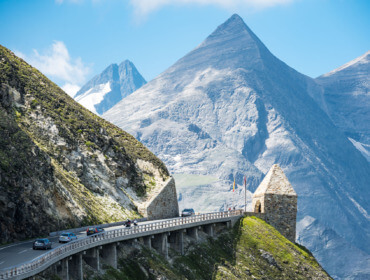 Auto's rijden over een kronkelende bergweg in het SalzburgerLand, omringd door stenen bogen en rotsachtige alpentoppen.