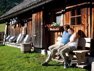 Een lachend stel zit op een bankje buiten een rustieke houten hut in de zon.