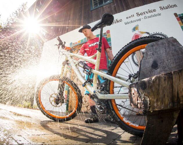 © SalzburgerLand Tourismus. f-bike-stories-saalachtal_beim_friedlwirt_25csalzburgerland_david-schultheiss-for-wom-medien Persoon wast een mountainbike met een spuitbus, zonlicht schijnt, bij een bord Bike Service Station.