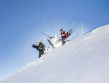Drie mensen met snowboards lopen bergopwaarts op een winterhelling onder een strakblauwe hemel, zonlicht achter hen.