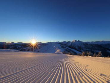 Zonsopgang boven besneeuwde Grossarltal bergen met geprepareerde skipistes en een strakblauwe lucht.