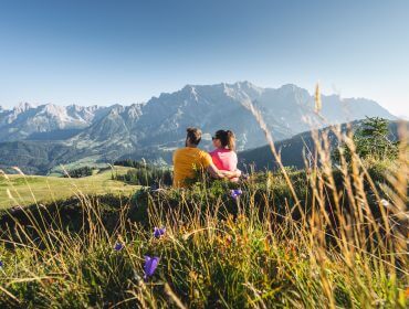 Een echtpaar zit op een met gras begroeide heuvel bij Hochkönig, uitkijkend over bergen onder een strakblauwe hemel en wilde bloemen.