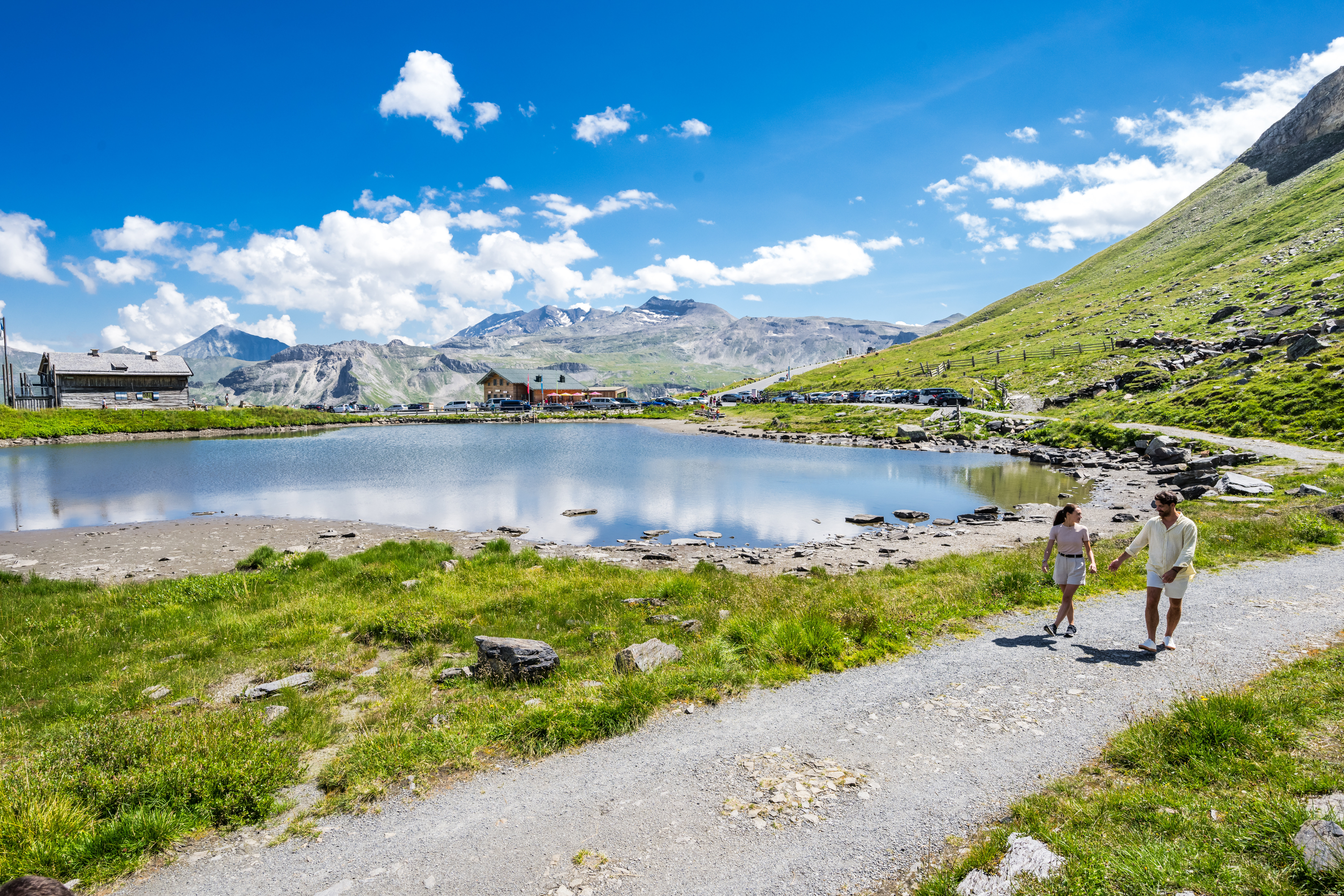 Twee mensen wandelen langs een bergmeer bij de Grossglockner Hochalpenstrasse, met alpentoppen op de achtergrond. (Vergrote weergave)