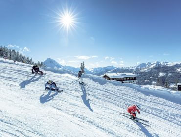 Vier mensen skiën in Wagrain-Kleinarl langs een besneeuwde helling onder een stralende zon met bergen op de achtergrond.