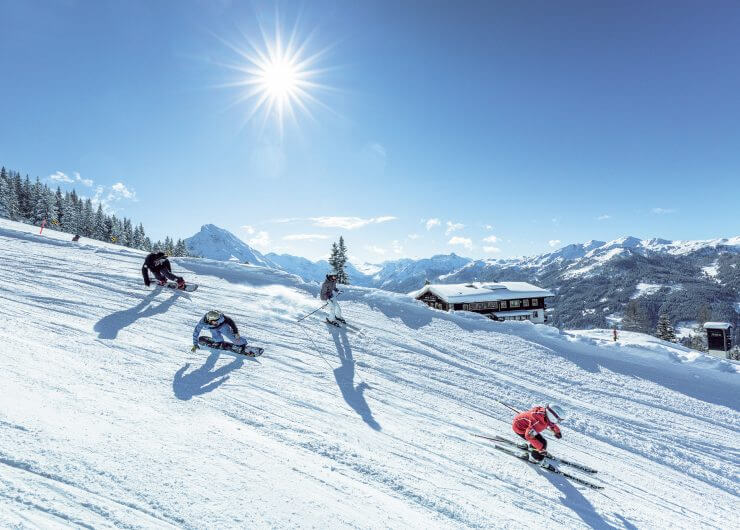 Cross(c)Shuttleberg Vier mensen skiën in Wagrain-Kleinarl langs een besneeuwde helling onder een stralende zon met bergen op de achtergrond.