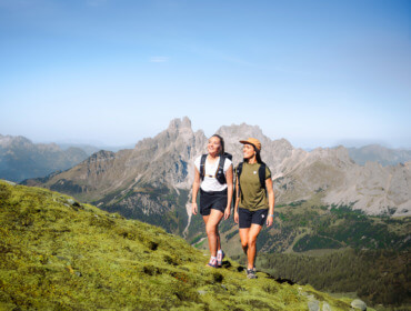 Twee mensen wandelen bergopwaarts in het SalzburgerLand, met gras begroeide berghellingen en rotsachtige pieken onder een blauwe hemel.