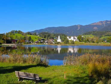 Een houten bank kijkt uit op het Goldegg meer met een dorp en bergen onder een helderblauwe hemel.