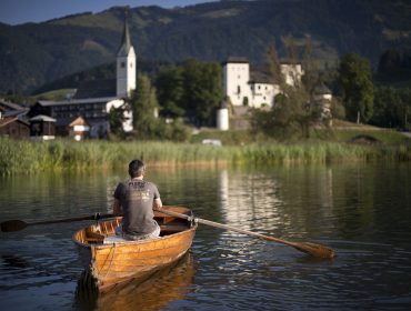 Een man roeit met een houten boot op het kalme meer van Goldegg, met een dorp en bergen op de achtergrond.