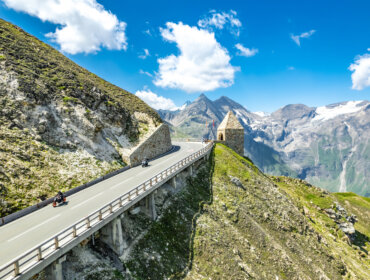 Motorrijders rijden over de Grossglockner Hochalpenstrasse, slingerend langs stenen muren en besneeuwde bergtoppen onder een blauwe hemel.